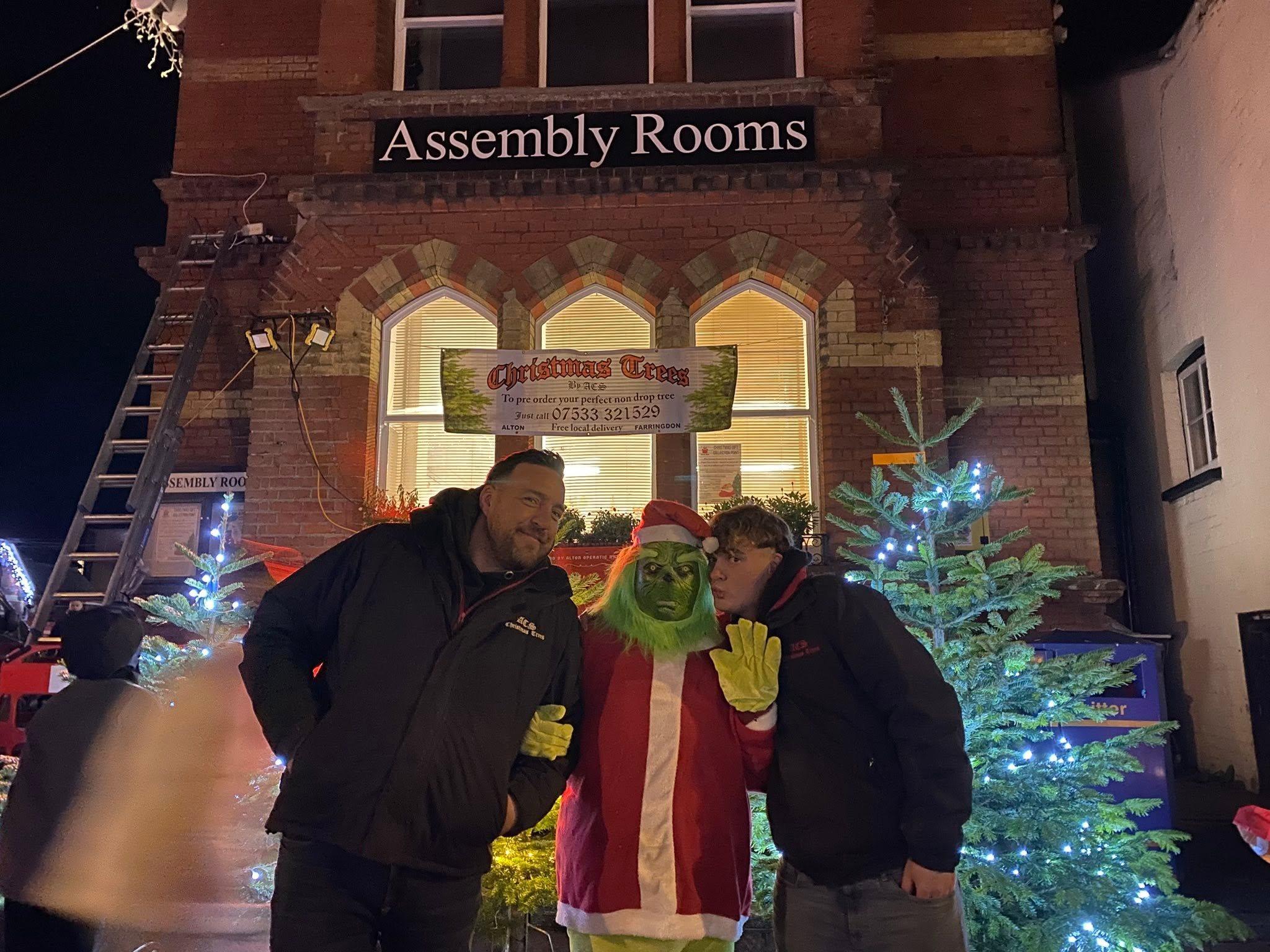 Two of the ACS team in branded jackets with the Grinch outside the Assembly Rooms at night